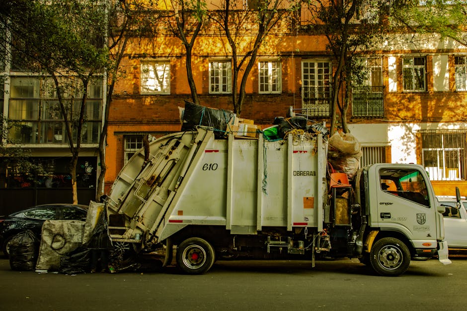 A black and white photograph of a commercial waste collection vehicle parked on a city street, with residential buildings and leafless trees in the background. The truck features a large, enclosed met