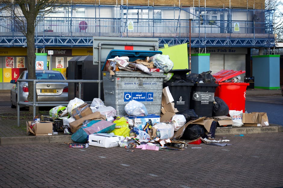 A sanitation worker wearing a high-visibility yellow and red uniform is standing behind a large red waste collection vehicle on a paved street, engaging in rubbish collection or street cleaning. The v